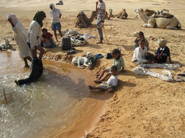 Children sitting on sand beside a small lake, men and camels nearby