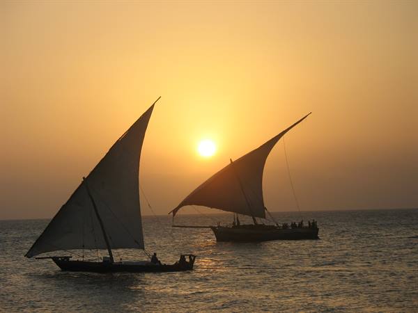 Two dhows with triangular sails glide past the low sun on the sea