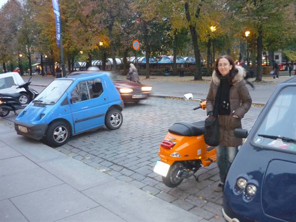 Woman beside orange scooter and blue microcar on cobblestones, Oslo