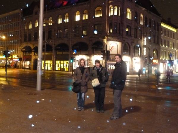 Three tourists pose in falling snow before illuminated buildings in Oslo at night