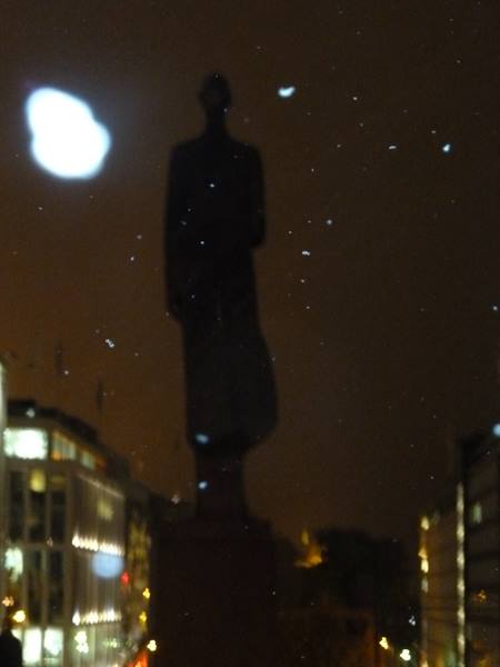 Standing statue at night in Oslo, soft glow, blurred lit buildings around