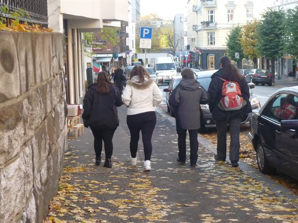 Pedestrians walk over autumn leaves on an Oslo sidewalk, stone wall on the left