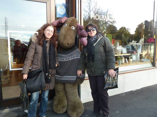 Two women with shopping bags beside a large stuffed moose outside a shop