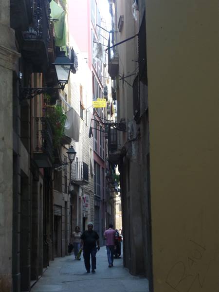 Pedestrians in a narrow alley between buildings with balconies, lamps and hanging laundry