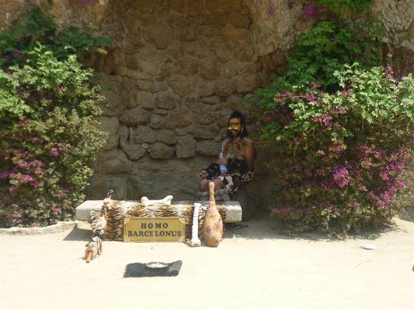 Painted man seated under stone wall with Homo Barcelona sign among flowers