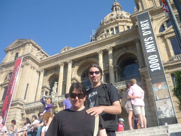 Two tourists before the Palau Nacional, dome and exhibition banners under sunny sky