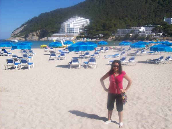 Woman in pink shirt on beach with blue umbrellas, loungers and hotel among pines
