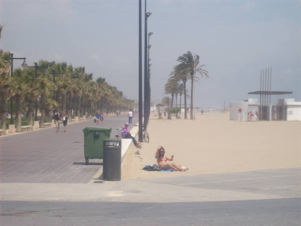 Woman sitting on sand by beach promenade with palm trees and walkers