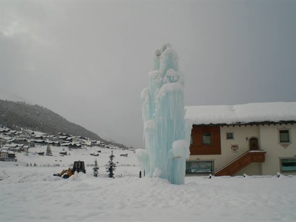 Blue ice fountain on snow-covered square next to wooden chalet