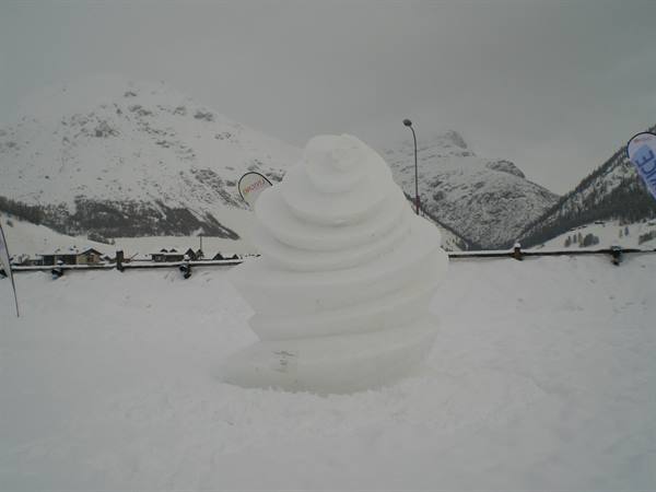 Spiral snow sculpture on snowy slope, mountains and cabins under grey sky