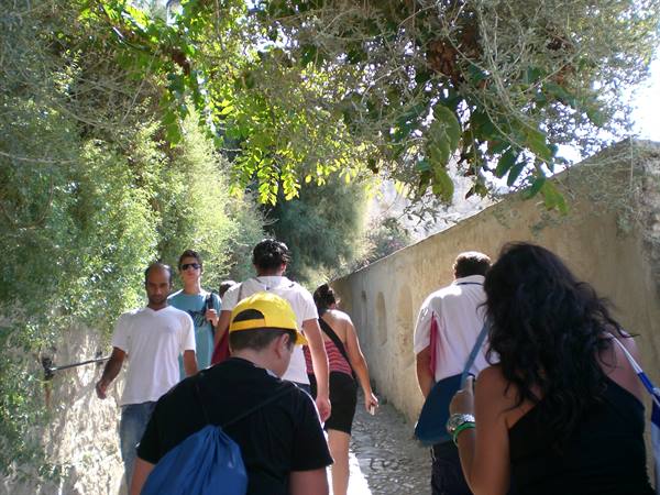 Tourists with backpacks walk along a tree-shaded path beside a stone wall
