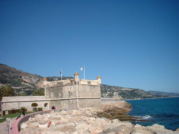 Stone bastion at Menton harbor, rocky shore and blue sea under clear sky
