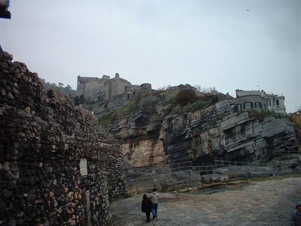 Two people walk on stone paving toward Portovenere castle under grey sky