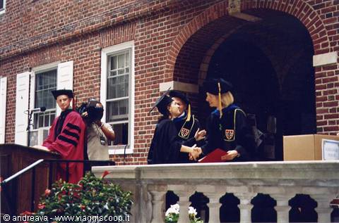 Graduates in black gowns and caps shake hands on a brick terrace
