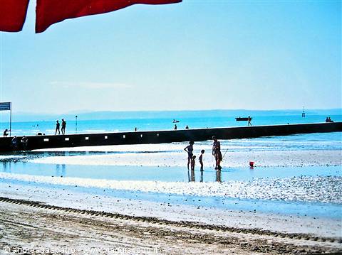 Bathers on shallow beach under red umbrella, breakwater stretching across the sea