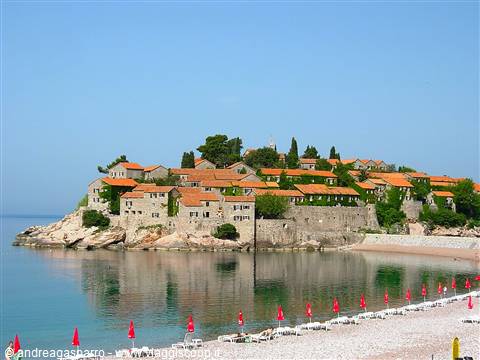Stone islet with orange-roofed houses reflected in calm sea, beach with red umbrellas