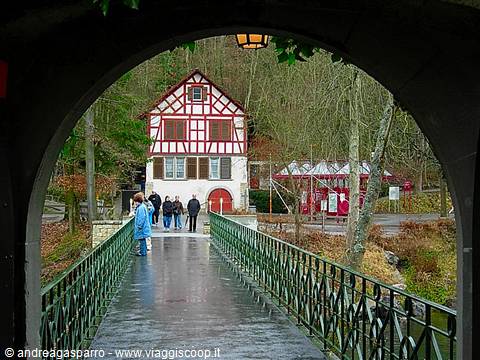 Half-timbered house framed by a stone bridge arch, people walking in the rain