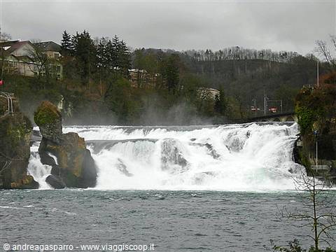 Powerful foamy waterfall between rocks, houses and bare trees under grey sky