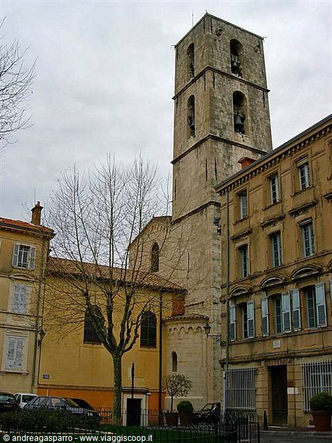 Stone bell tower among ochre buildings, grey sky, bare trees, parked cars