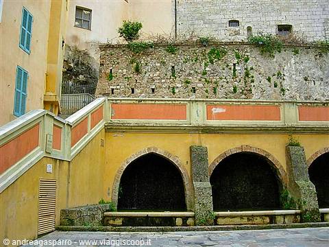 Old arched fountain with stone basins, yellow facade and plant-covered wall
