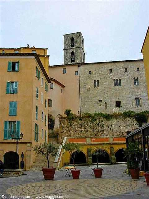 Stone bell tower above ochre houses, square with potted olive trees