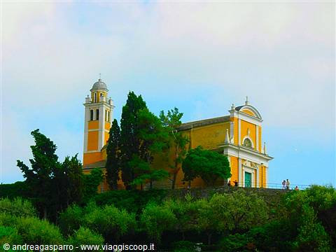 Yellow church with bell tower among green trees under cloudy sky