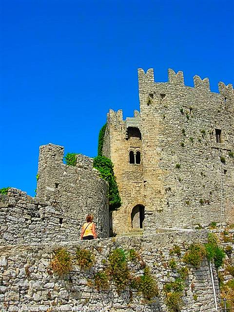 Woman walks toward the crenellated stone tower of Erice castle, bright sunlight
