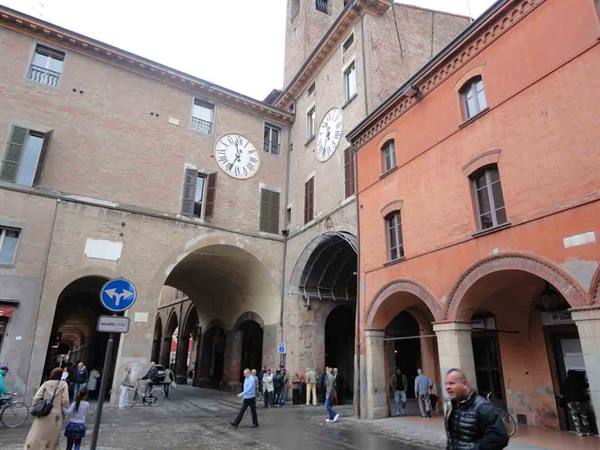 Clocks on brick buildings above arcades, pedestrians crossing the wet square