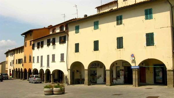 Square with ochre arcaded buildings, green shutters, parked cars and stone planters