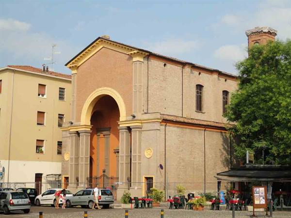 Brick church facade with large arch, square with parked cars and café tables