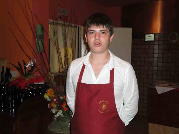 Young waiter in red apron among bottles and flowers inside the restaurant