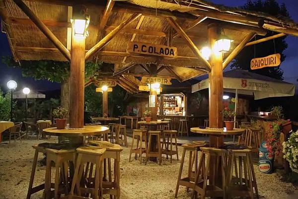Open-air wooden thatched bar with stools and lit Piña Colada signs at night