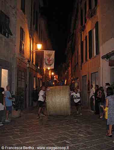 Men push a giant cheese wheel down the lamplit alley, crowd watching