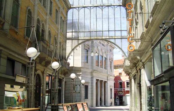 Glass-covered arcade with Ottico sign, historic buildings and globe lamps, Trieste