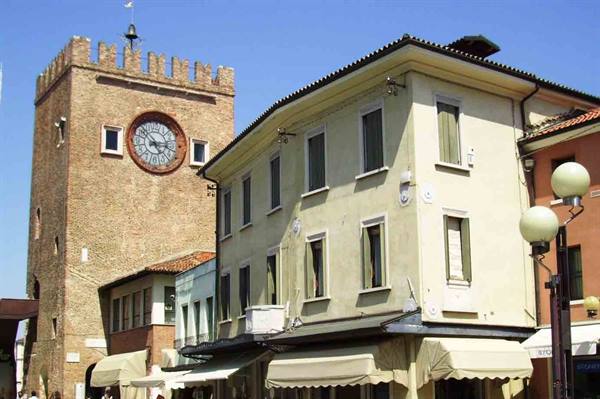 Brick crenellated clock tower beside pastel buildings with awnings under blue sky