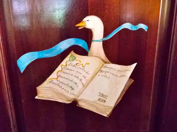 Painting on wood: a white goose with blue ribbon leafs through an old book