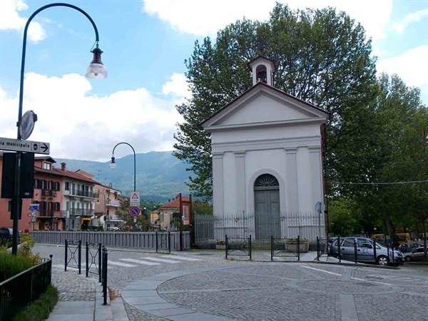 Small white chapel with bell tower at a crossroads, mountains and houses behind
