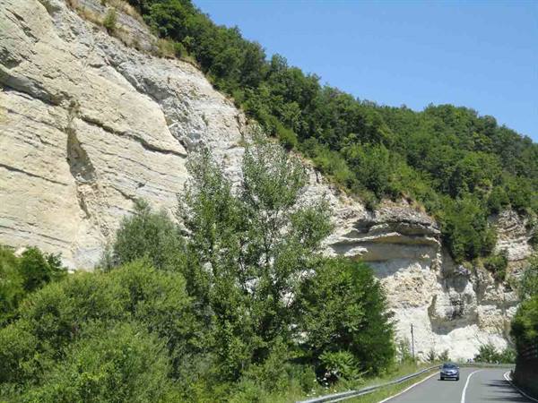 Car on asphalt road beneath pale rock cliffs with green trees, blue sky