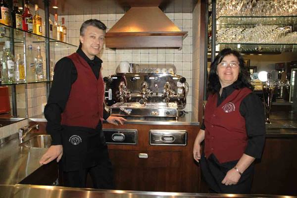 Two bartenders in red vests behind a chrome coffee machine