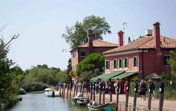 Tourists along Torcello canal, red houses, moored boats and brick bridge