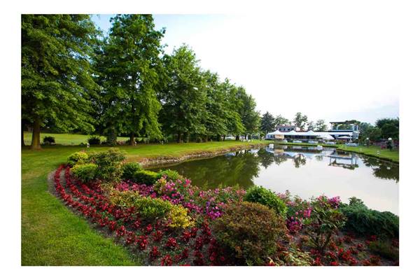 Pond with red and pink flowers, tall trees reflected in water, white pavilion