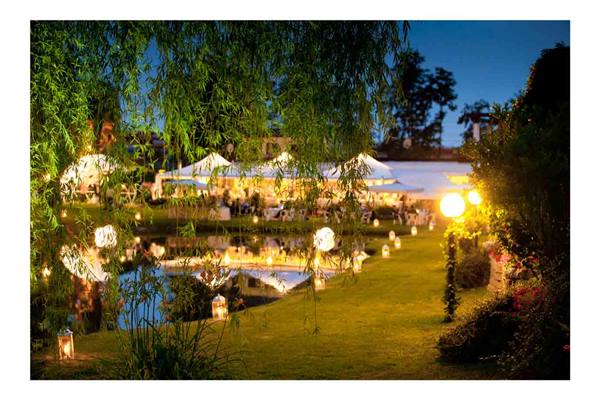 Lit lanterns along the pond, glowing tents between willow and lawn