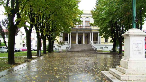 Rain-soaked tree-lined avenue leading to neoclassical villa with staircase, marble monument on right