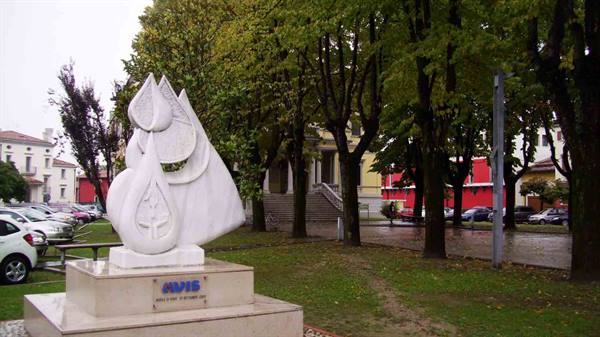 White marble droplet sculpture in tree-lined square, red building behind