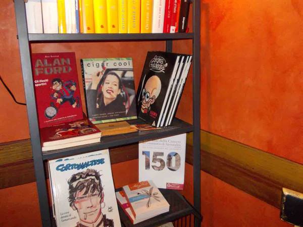 Metal shelf with books and Corto Maltese comics against orange wall, warm light