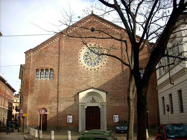 Brick church facade with rose window, bare tree branches, parked cars and cloudy sky