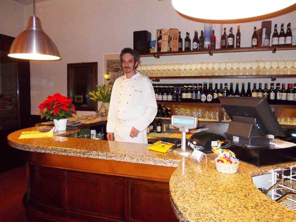 Chef in white behind granite bar counter, wine bottles and metal lamps