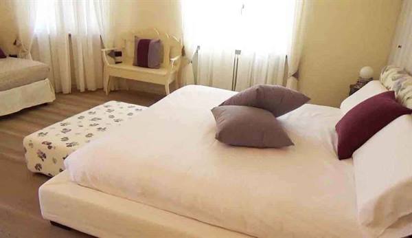 Bedroom with white bed, grey and burgundy pillows, bright sheer curtains