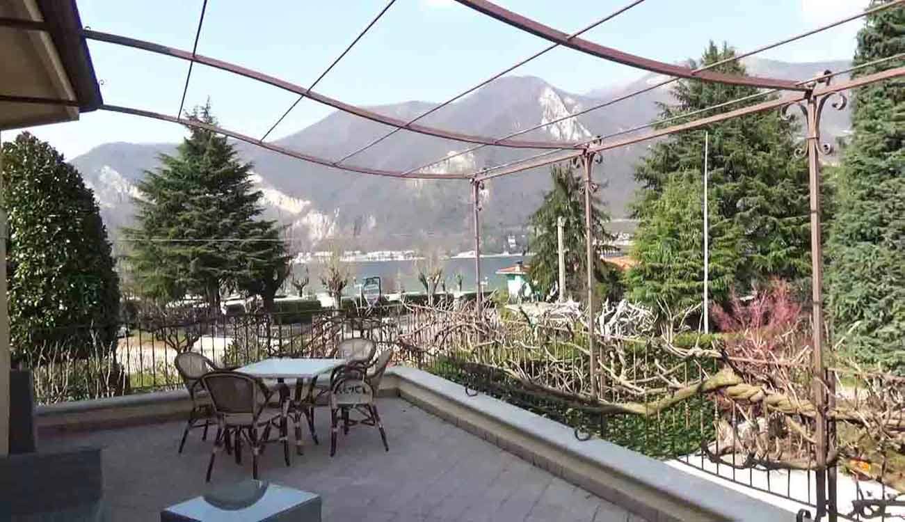 Tavolo con sedie sotto pergolato di ferro sul lago, montagne sullo sfondo