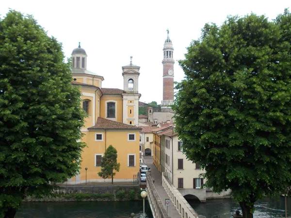 Yellow church and clock tower of Bassano del Grappa among green trees and bridge